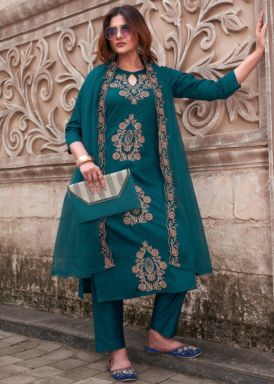 Woman in teal embroidered outfit standing against a decorative wall.