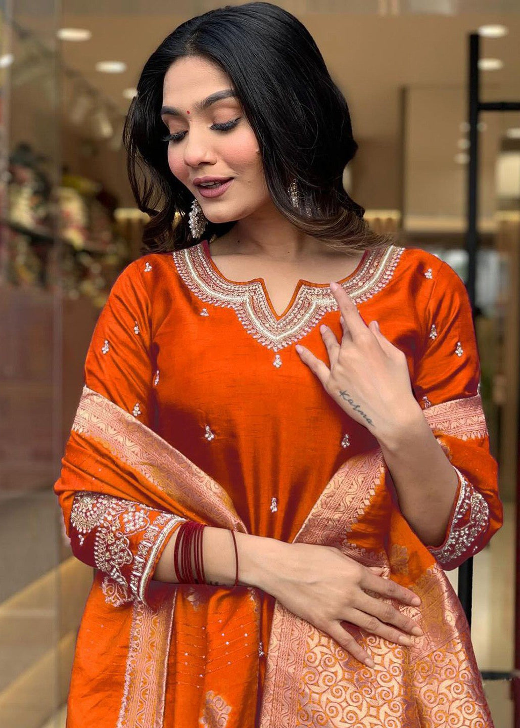 Woman wearing an orange traditional outfit with intricate patterns indoors.