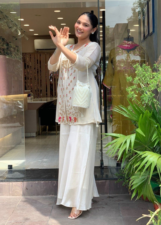 Woman wearing a vibrant orange Roman Silk embroidered kurti paired with a matching flared sharara featuring elegant white floral embroidery and a V-neck design, ideal for festive and ethnic occasions.
