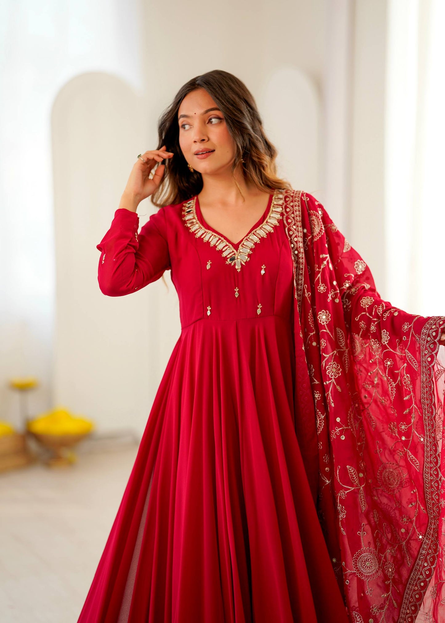 A woman modeling a long-sleeved red Anarkali gown with gold embroidery on the neckline and a matching red embroidered dupatta, posed in a bright, minimalist indoor setting.