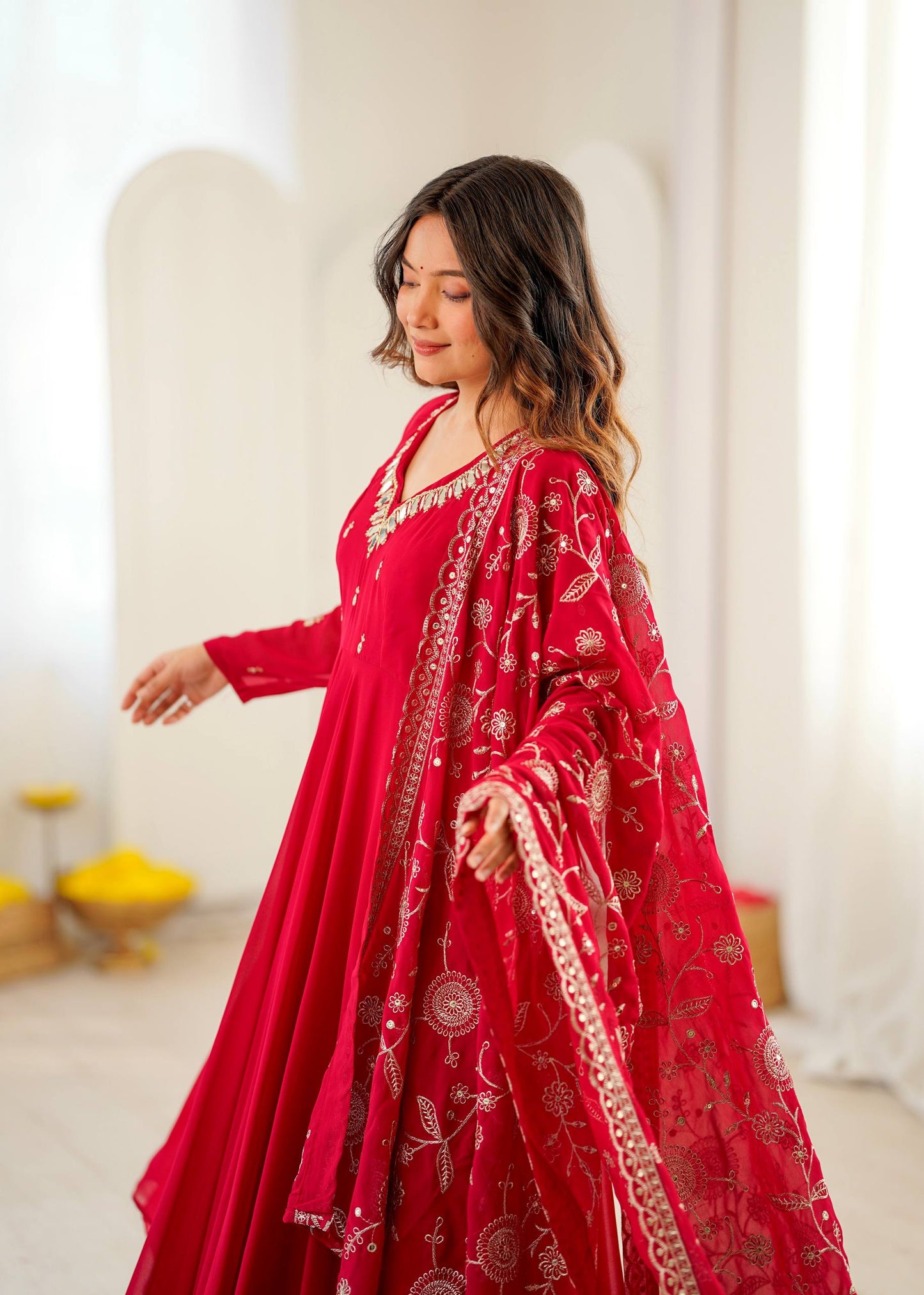 A woman wearing a vibrant red traditional outfit with intricate gold embroidery, standing in a bright, softly lit indoor setting with white arches in the background.