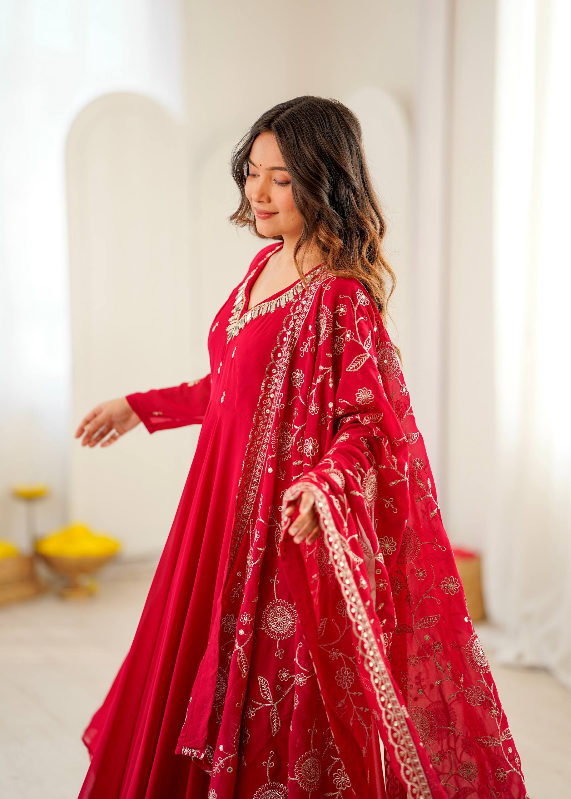 A woman wearing a vibrant red traditional outfit with intricate gold embroidery, standing in a bright, softly lit indoor setting with white arches in the background.