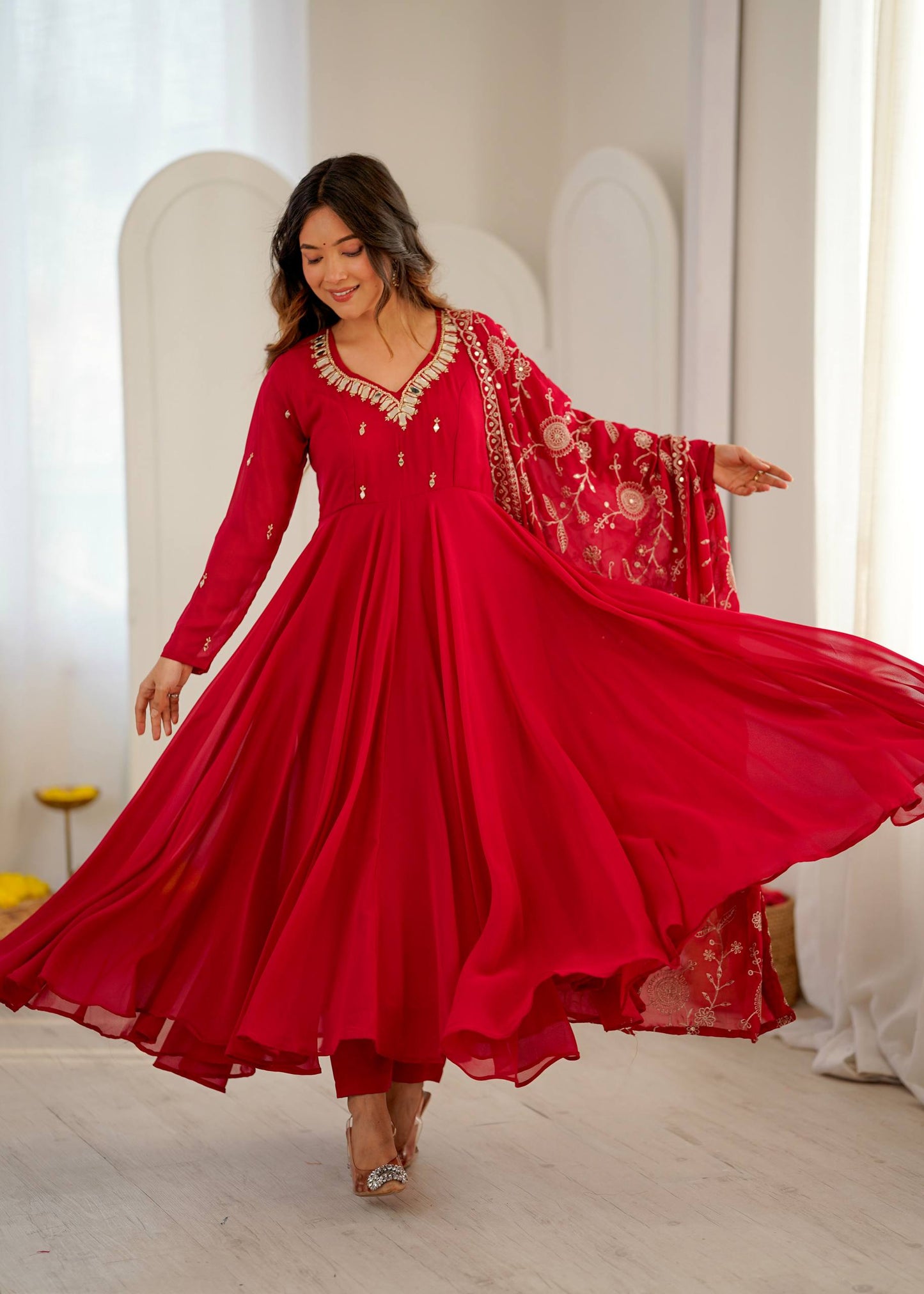 A woman wearing a flowing red long kurta with gold embroidery and a matching embroidered dupatta, captured mid-twist in a bright indoor setting with white arches and curtains.