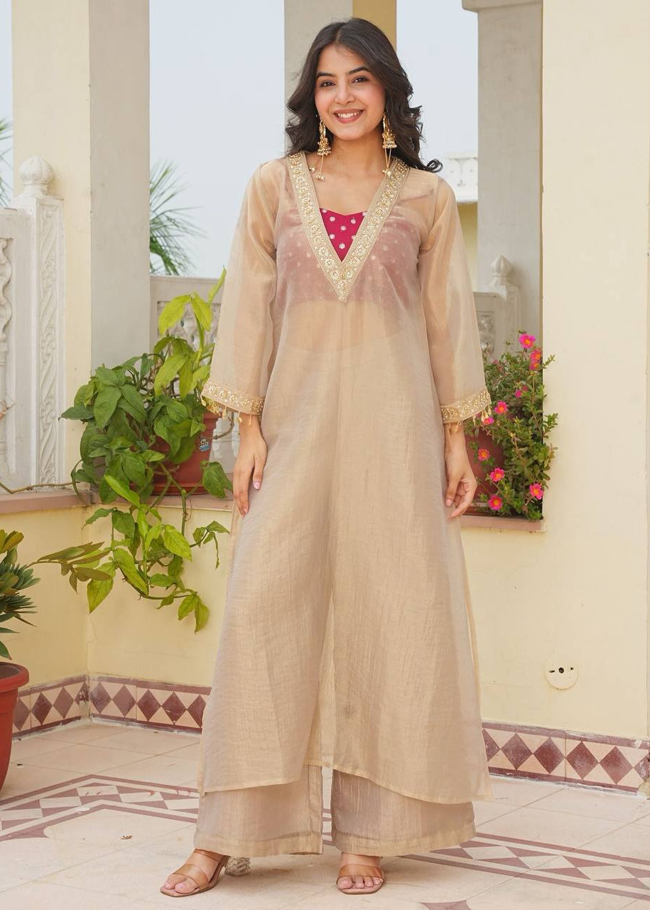A woman modeling a beige sheer kurta with gold embroidery and matching wide-leg trousers, styled with a pink polka-dot top, in an outdoor patio setting.