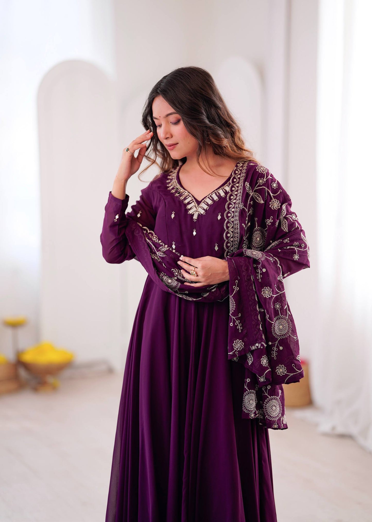 A woman wearing a long-sleeved deep purple maxi dress with silver embroidery and a matching embroidered shawl, standing in a bright, minimalist indoor setting.