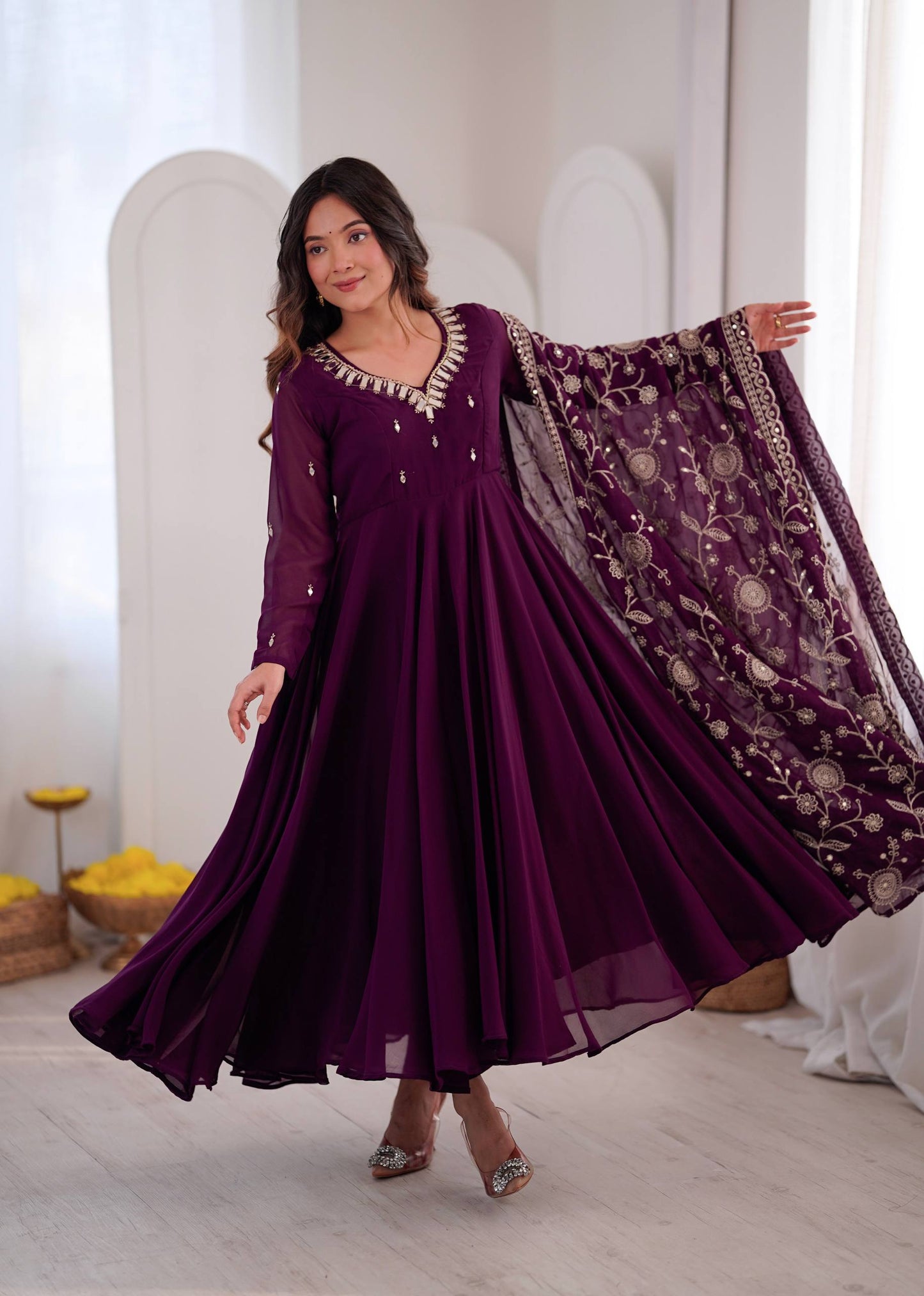 A woman models a deep purple long-sleeve Anarkali gown with silver embroidery on the neckline, paired with a matching sheer embroidered dupatta, in a bright indoor setting.