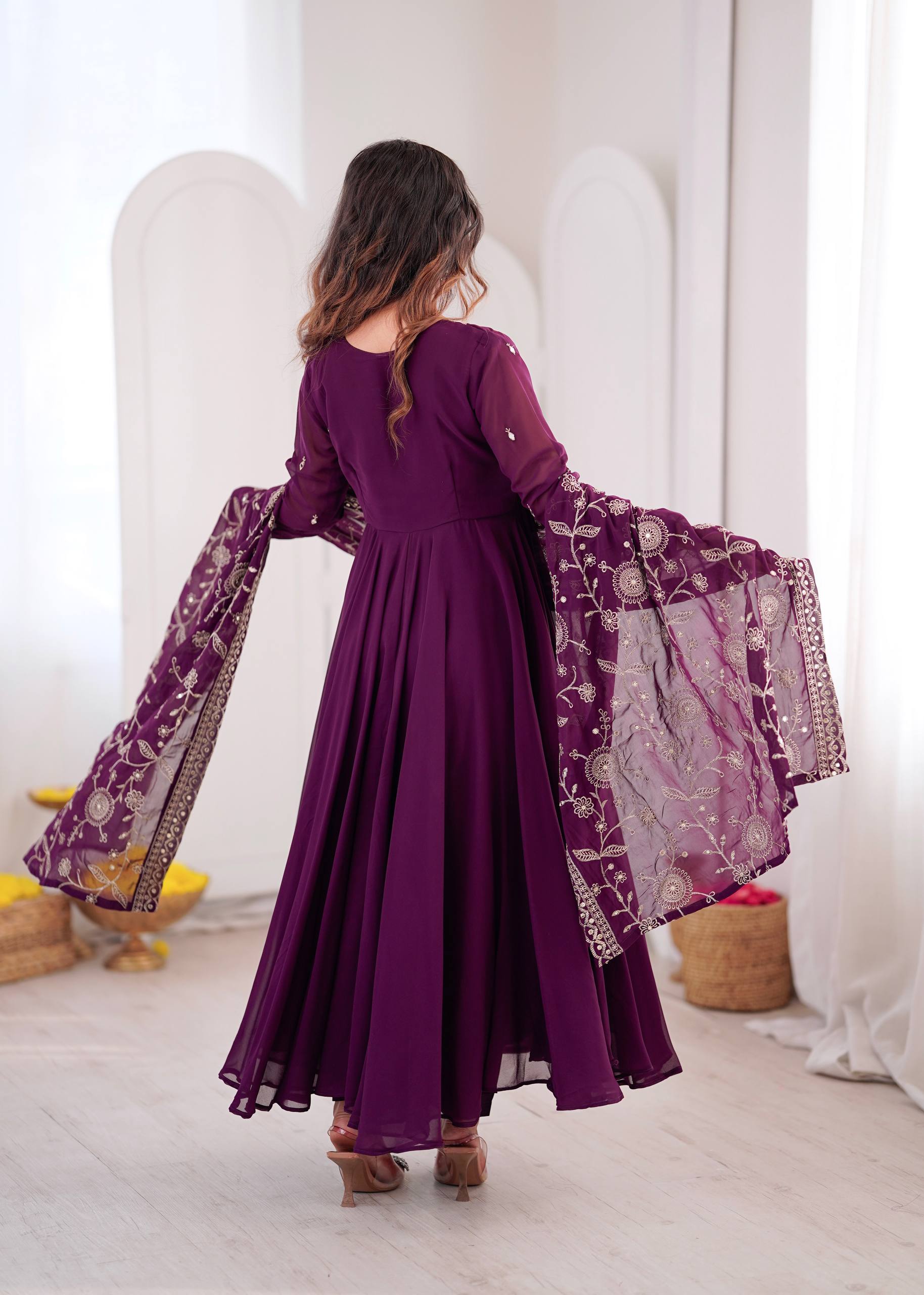 A woman wearing a deep purple long-sleeve maxi dress with a matching sheer embroidered dupatta, shown from the back in a bright, modern indoor setting.