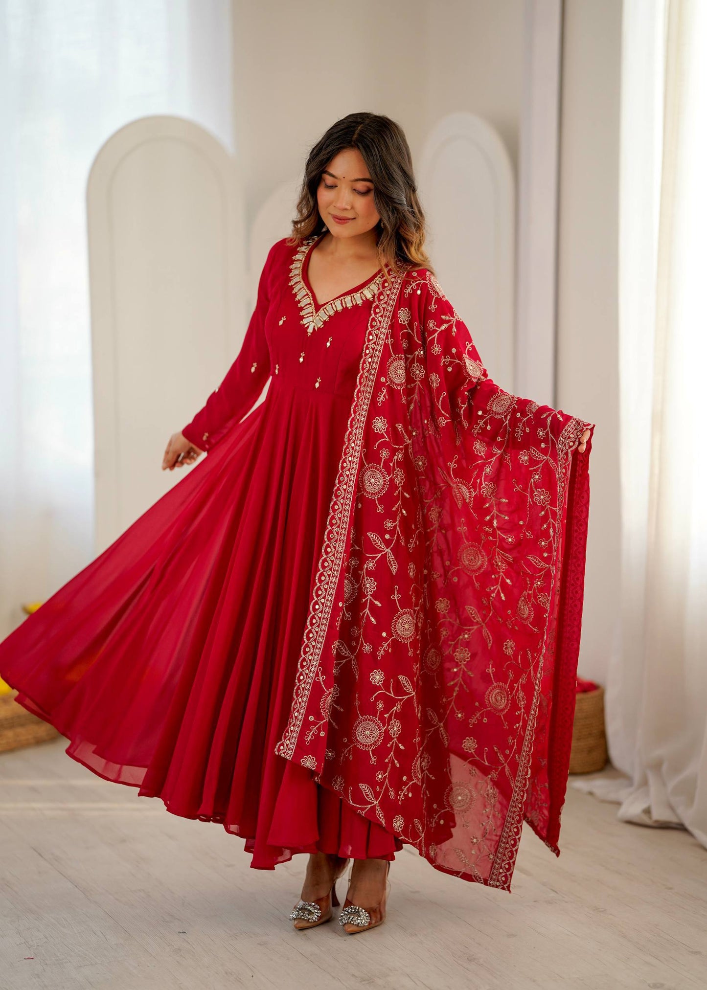 A woman modeling a vibrant red traditional Indian outfit with a matching sheer dupatta, decorated with intricate gold and silver embroidery, set against a bright, minimalist indoor background.
