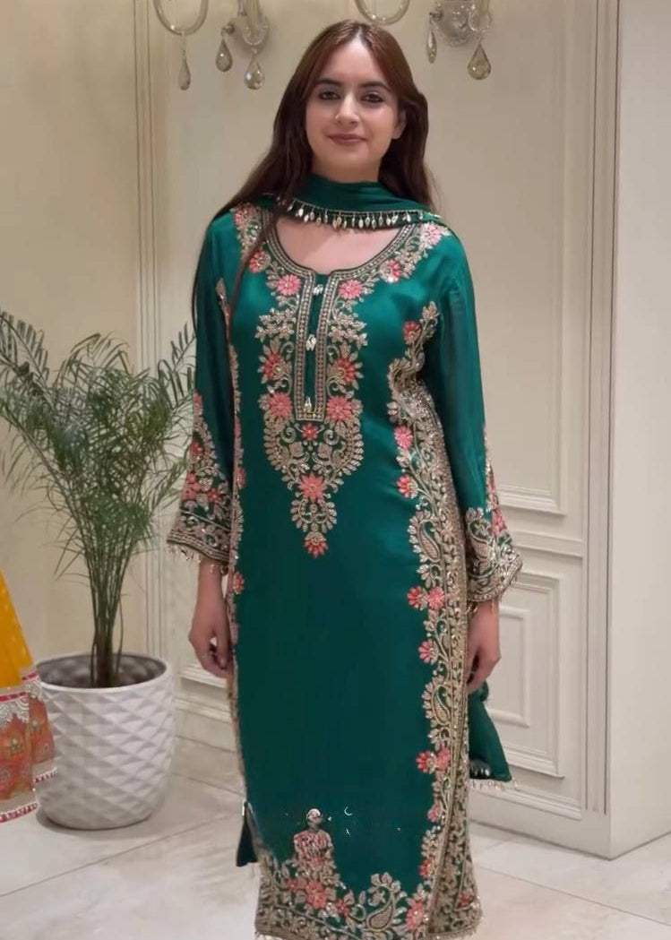 A woman modeling a traditional green salwar kameez suit with intricate gold and pink floral embroidery, standing in an indoor setting with a potted plant and wall sconces in the background.