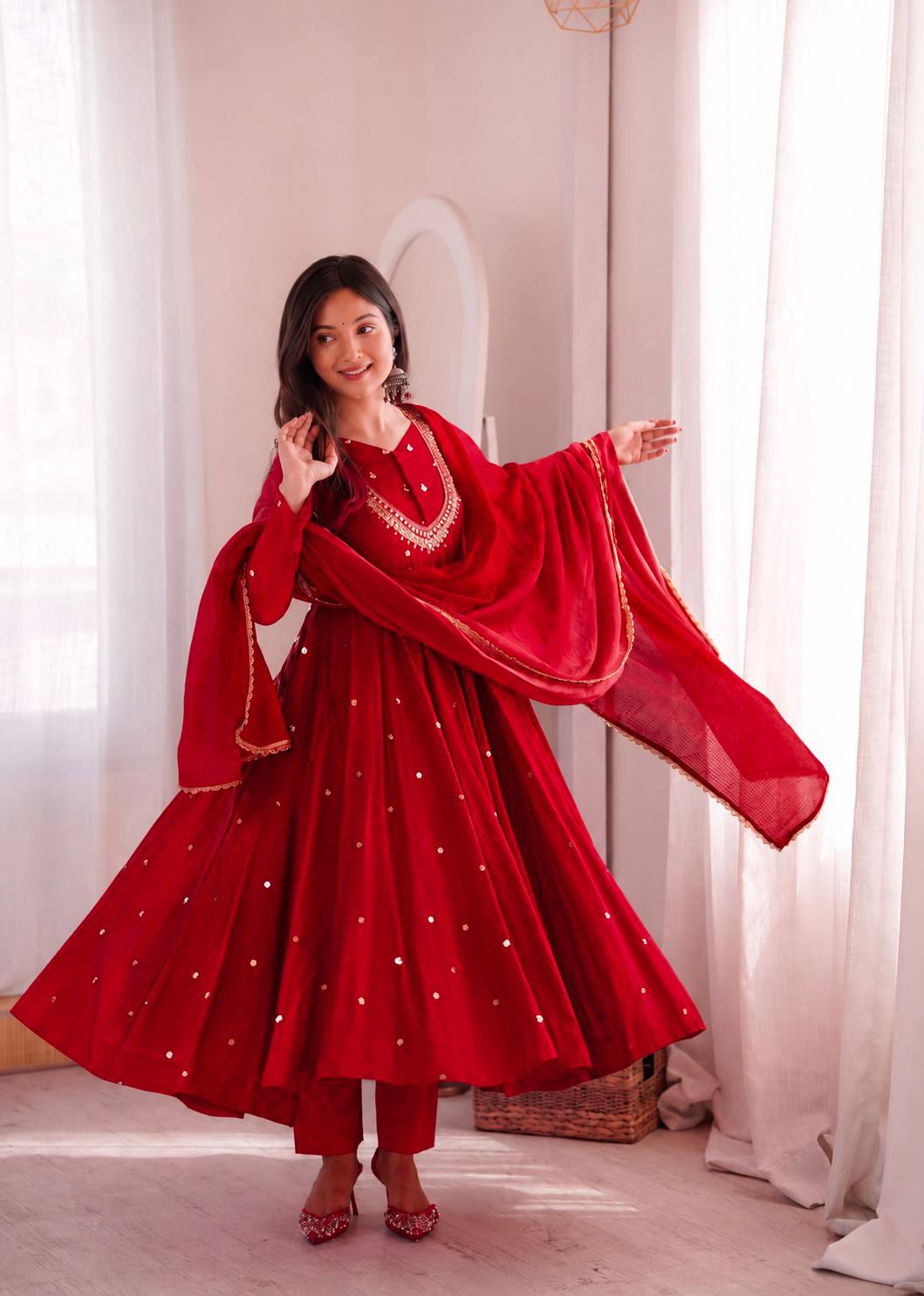 A woman modeling a traditional red Indian salwar kameez outfit with a matching dupatta, featuring gold embroidery and silver mirror work, in a softly lit room with white curtains.