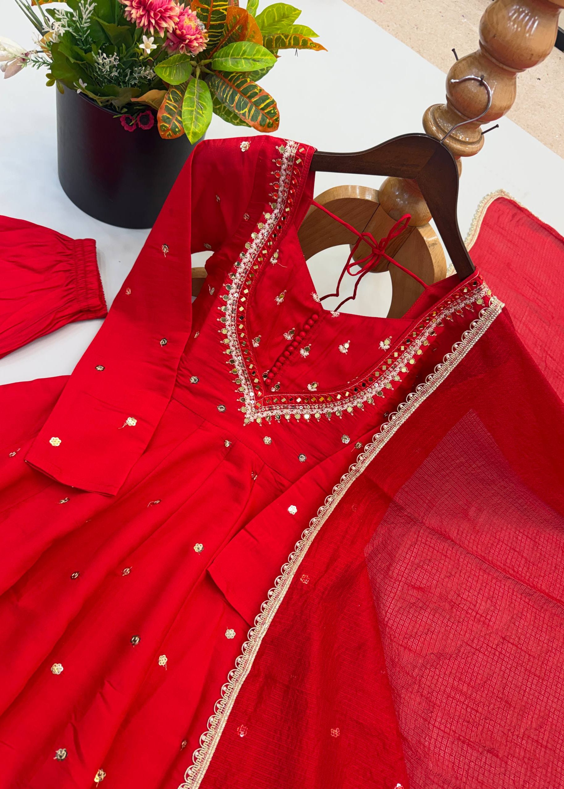 A bright red traditional dress with gold embroidered details and a matching sheer red dupatta draped over a wooden hanger, with a matching red sleeve laid to the left on a white surface.