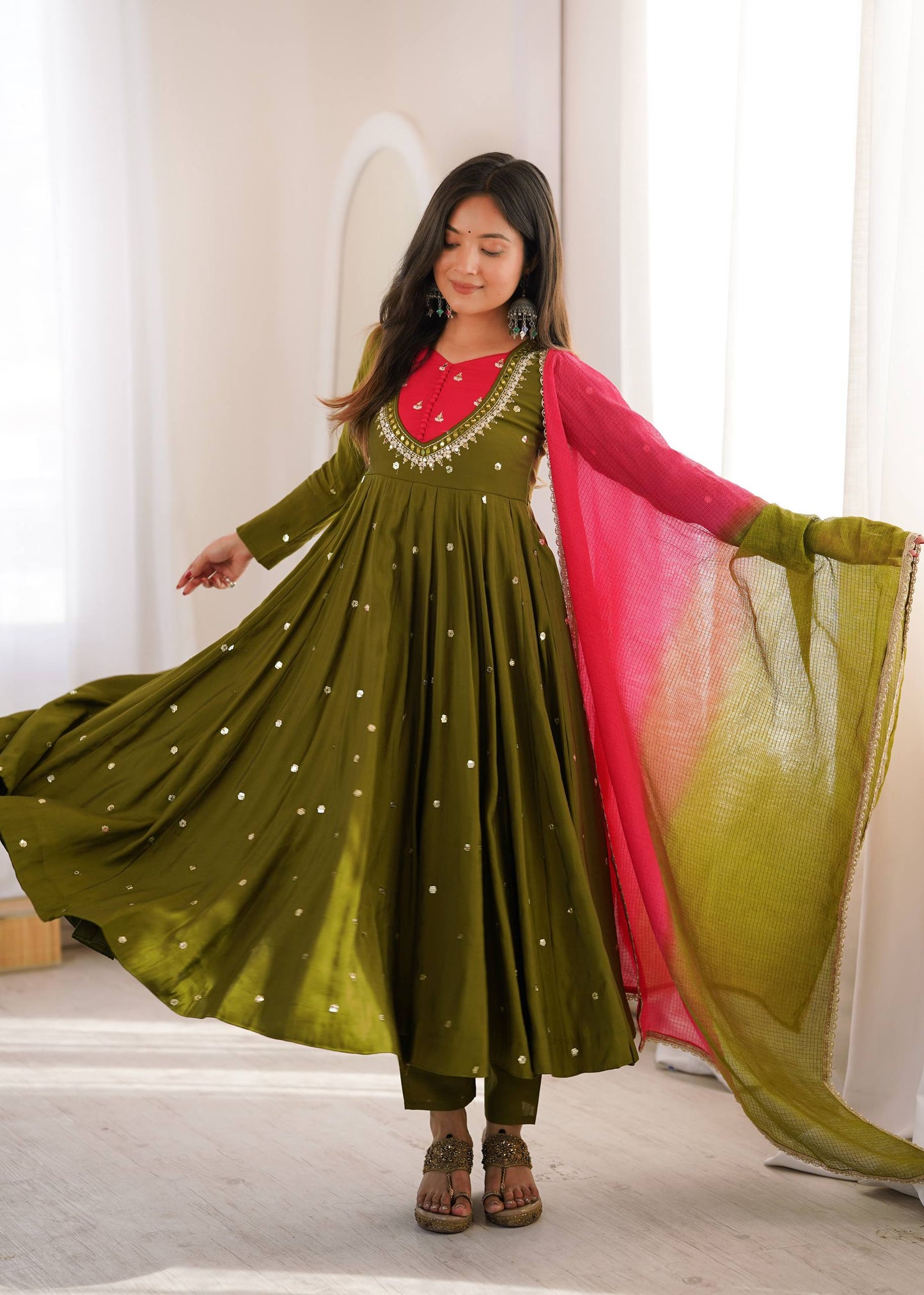 A woman modeling a traditional olive green Anarkali gown with gold embroidery, a bright pink inner lining, and a matching pink-to-olive gradient dupatta, set against a bright, minimalist indoor background.