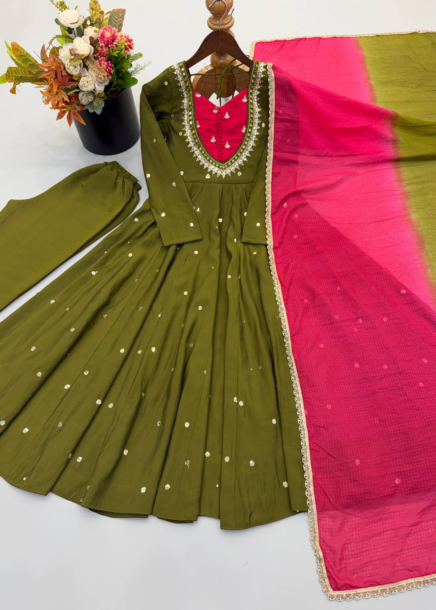 An olive green Anarkali gown with a bright pink and olive green dupatta, laid out on a white surface next to a pair of matching olive green trousers. The dress features a red embroidered neckline with gold beadwork and small white embroidered dots.