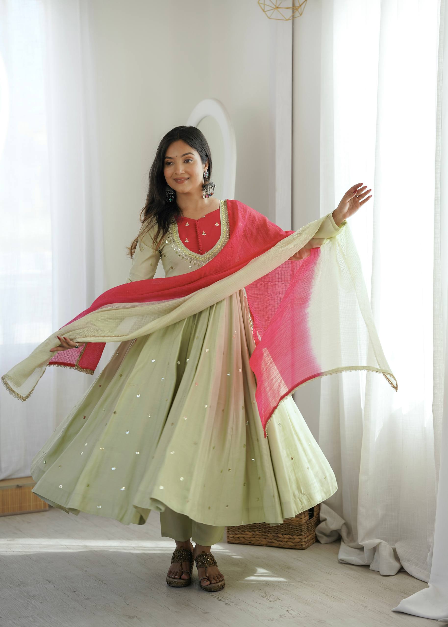 A woman modeling a traditional Indian outfit consisting of a light green and pink gradient anarkali skirt, a red embroidered bodice, and a bright pink dupatta, set against a bright room with white curtains.