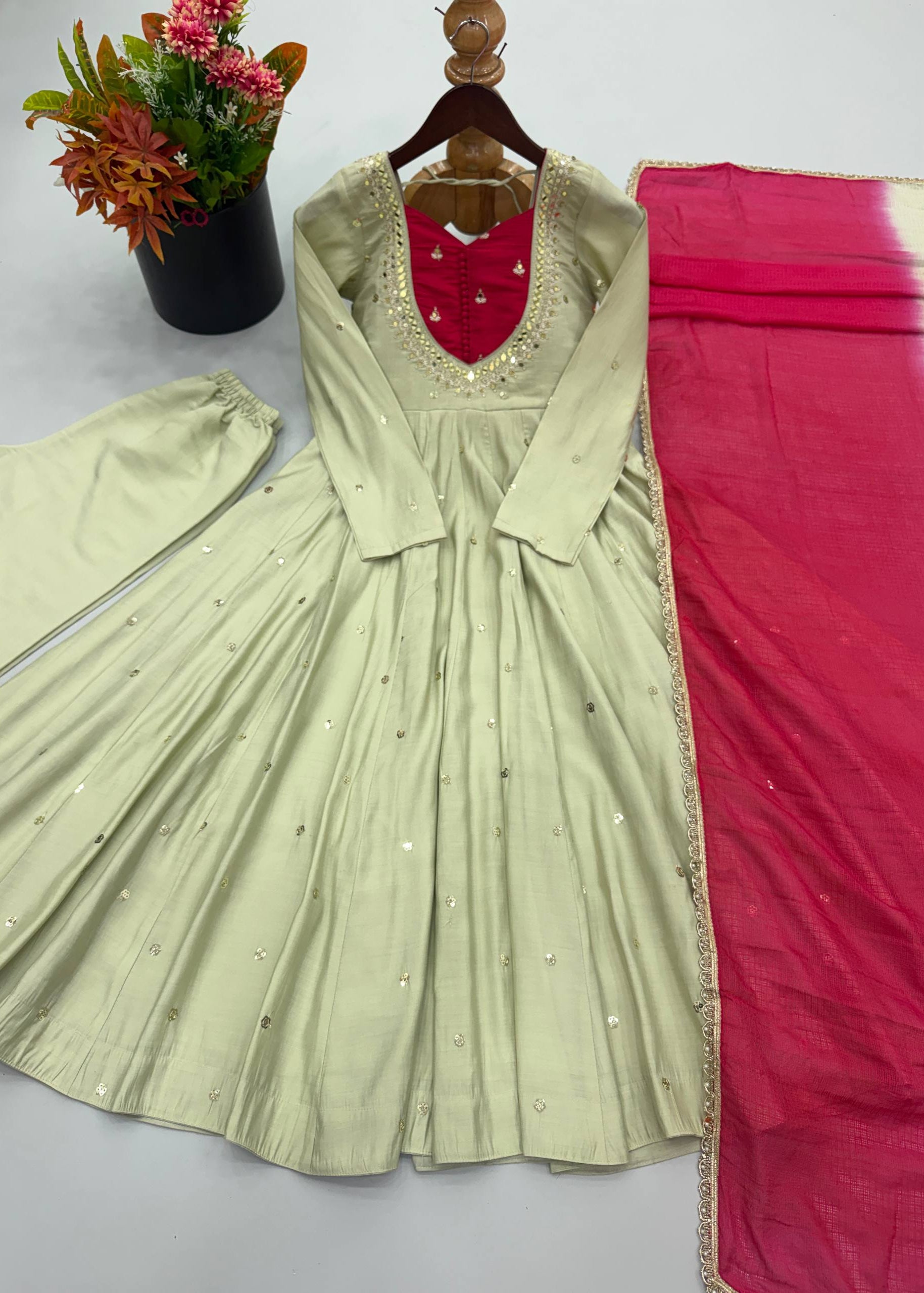 A light sage green Anarkali gown with silver mirror work embellishments, a red embroidered neckline, and a bright pink dupatta, displayed on a wooden hanger with a matching green pleated skirt piece and a floral pot in the background.