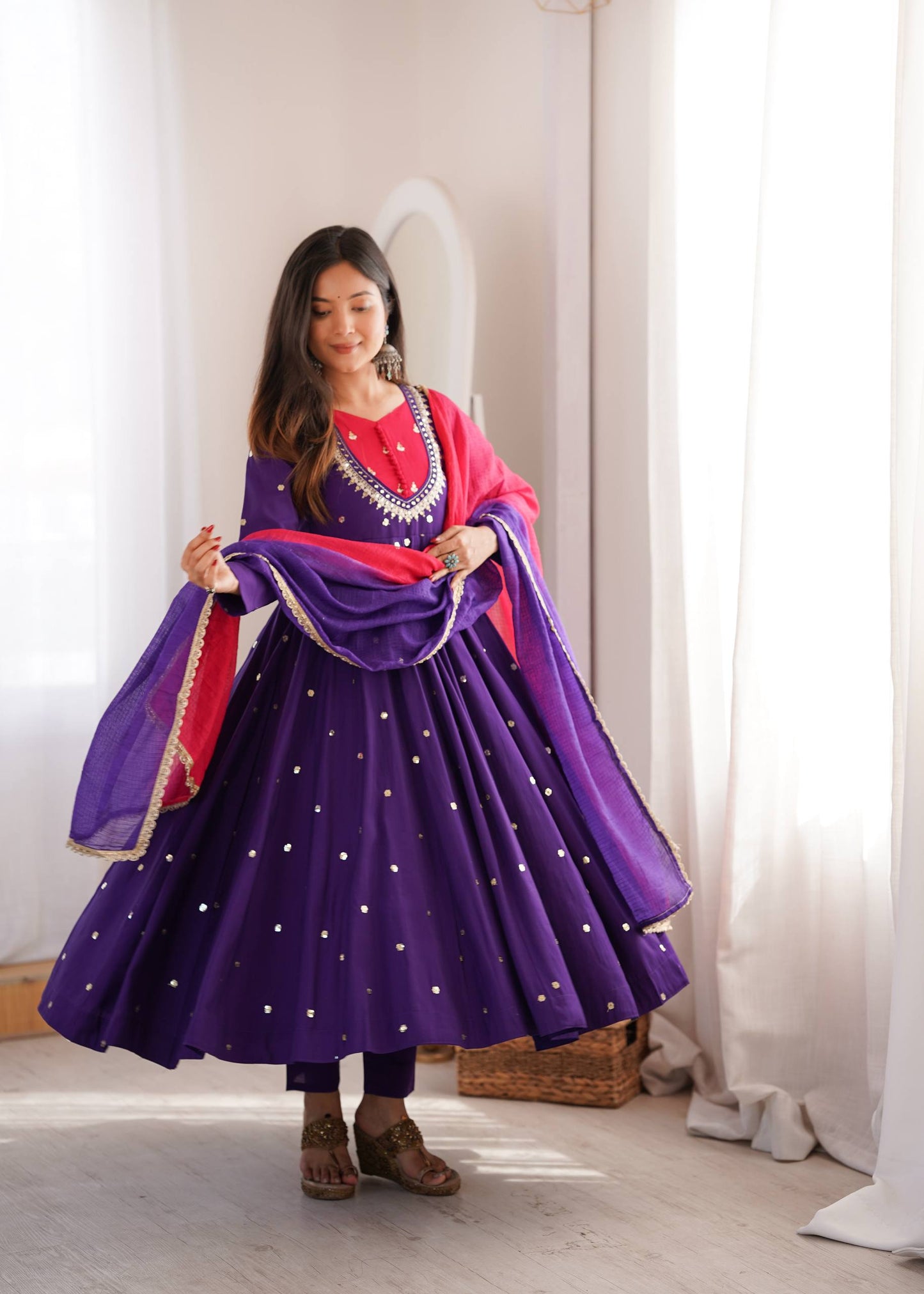 A woman modeling a traditional Indian outfit consisting of a purple flared skirt with gold star patterns, a pink top, and a purple dupatta with red and gold borders, set against a bright, softly lit room with white curtains.