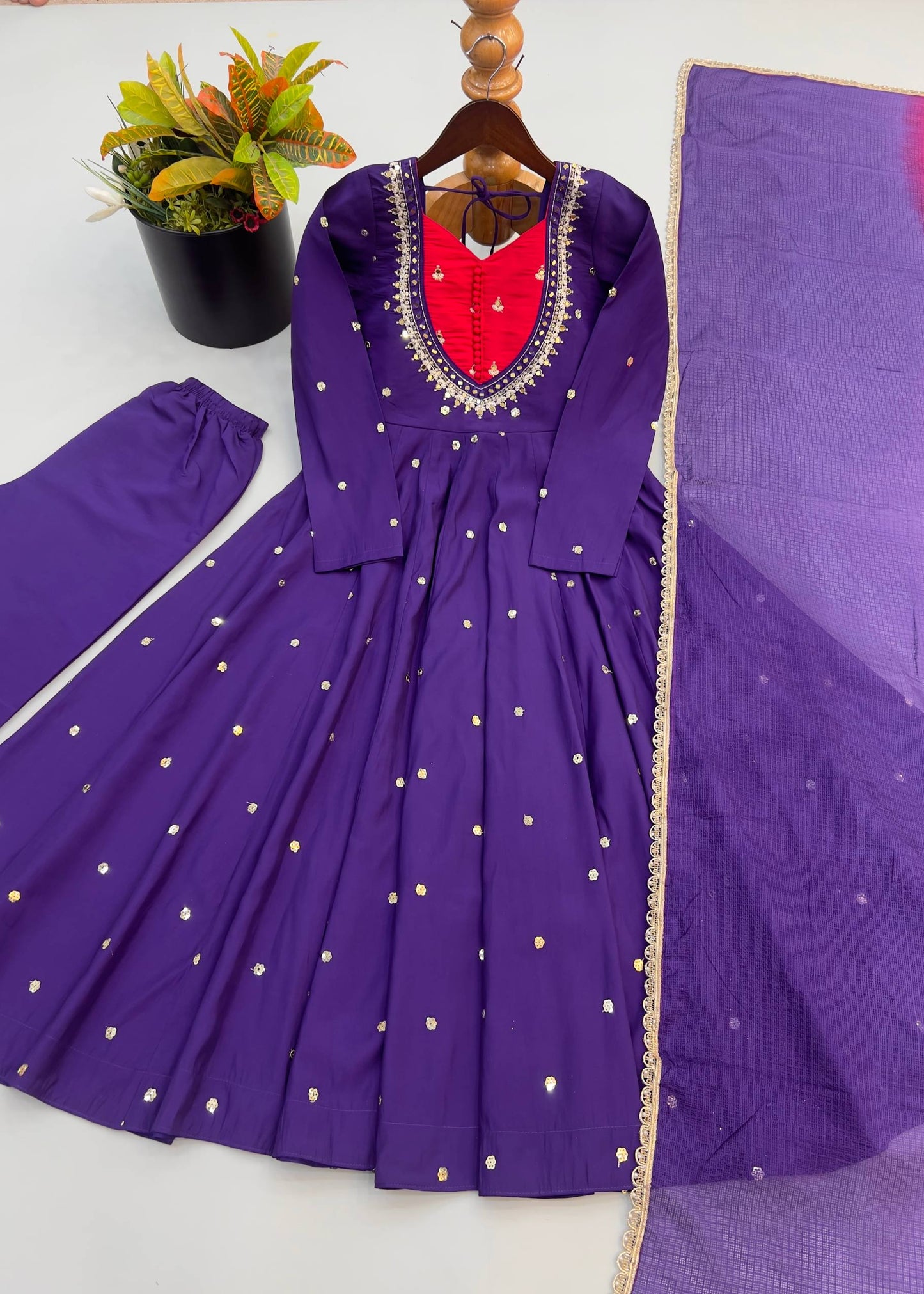 Purple long-sleeve Anarkali gown with gold dot embellishments, a red inner lining at the neckline, and a matching purple dupatta, displayed on a hanger with a plant prop on a white surface.