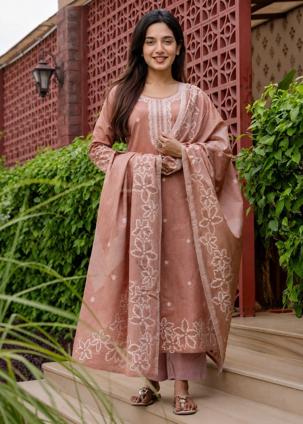 Woman in a traditional outfit standing outdoors with greenery and a decorative wall in the background
